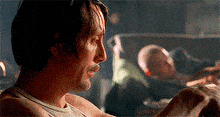a man without a shirt is sitting in a dark room with a man laying in the background .