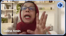 a woman wearing glasses and a red hijab is talking on a video call with salina amin .