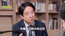 a man in a suit and tie is talking into a microphone in front of a bookshelf in a library .