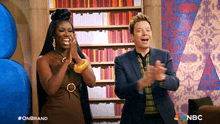 a man and a woman are clapping in front of a bookshelf with the nbc logo on the bottom