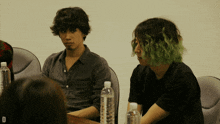 a man with green hair sits at a table with two bottles of water with the letter d in the corner