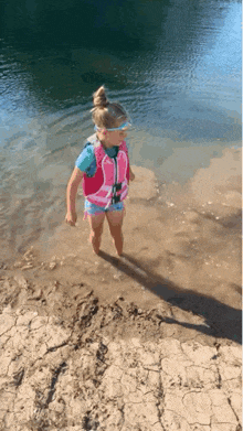 a little girl wearing a life jacket is standing in the water near a lake .