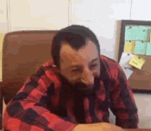 a man with a beard is sitting at a desk wearing a plaid shirt .