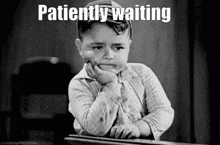 a little boy is sitting at a desk with his hand on his chin and the words patiently waiting written above him .