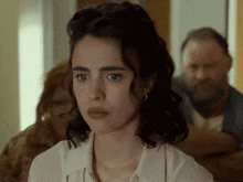 a woman in a white shirt looks at the camera with a man in the background