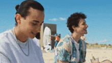 two men are sitting on a beach and one of them is wearing a hawaiian shirt