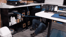 a person is kneeling under a desk with a laptop and a trash can