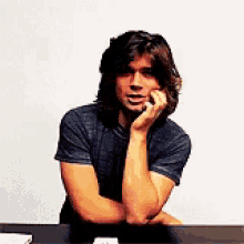 a man with long hair is sitting at a table with his hands on his chin