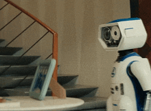 a white and blue robot is standing in front of stairs and a table