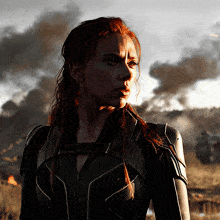 a woman in a black suit stands in a field with smoke in the background