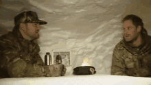 two men in military uniforms are sitting at a table and one of them has a beret with the word army on it