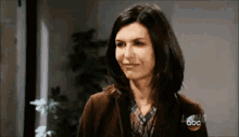 a woman in a brown jacket and plaid shirt is smiling in front of a plant .