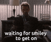 a man in a suit and tie is sitting in front of a sign that says " waiting for smiley to get on "
