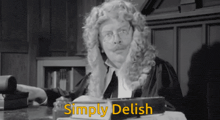 a man in a wig is sitting at a desk with the words simply delish written in yellow