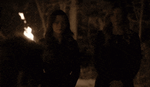 a woman in a black jacket stands in front of a fire