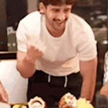 a man in a white t-shirt is standing in front of a table with food on it .