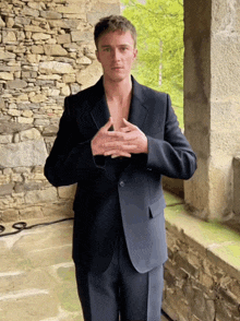 a man in a suit stands in front of a stone wall with his hands on his chest