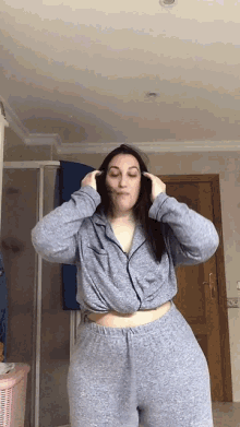 a woman in a grey pajama set is standing in a bathroom with her hands on her head