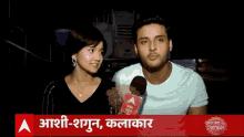 a man and a woman are talking into a microphone in front of a sign that says aashi