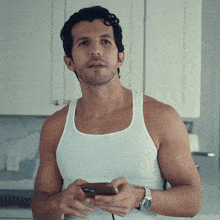 a man wearing a white tank top is holding a cell phone