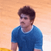 a man with a mustache is wearing a blue shirt and sitting on a wooden floor .