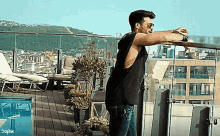 a man in a black tank top and sunglasses is leaning on a railing on a balcony overlooking a city .