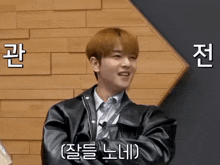a man wearing a black leather jacket is smiling in front of a wooden wall with korean writing on it
