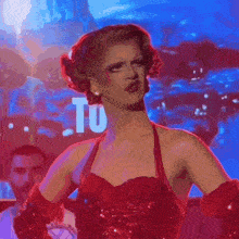 a drag queen in a red dress stands in front of a sign that says " tu "