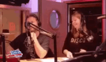 a man and a woman are talking into microphones in a studio . the man is wearing a shirt that says disco .