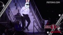 a man wearing sunglasses and a bunny head is dancing on a stage with pistons.com written on the bottom