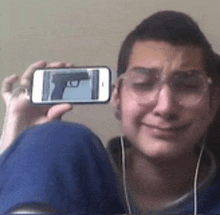 a man wearing glasses is taking a picture of himself with a gun on his phone .