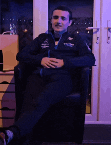 a man wearing a jacket that says williams racing is sitting in a chair