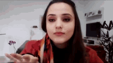 a woman in a red jacket and scarf is sitting in a living room with her hand out .
