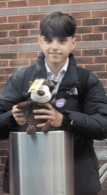 a boy wearing a north face jacket holds a stuffed animal in front of a brick wall