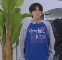 a young man wearing a blue sweatshirt that says `` very close , not yet '' is standing in front of a plant .