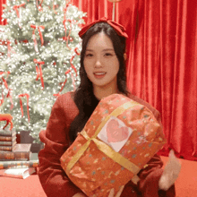 a woman in a red sweater is holding a gift with a heart on it in front of a christmas tree