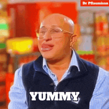 a bald man wearing glasses and a vest says yummy with his tongue out