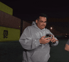 a man in a grey sweatshirt looks at his phone in front of a yellow container that says msc