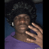 a man wearing a black hat and a purple shirt has his eyes closed and his hand on his face