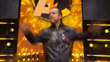 a man in a leather jacket is standing on a stage with his arms outstretched in front of a yellow background .