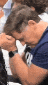 a man in a blue shirt is covering his face with his hands while sitting in a crowd of people .