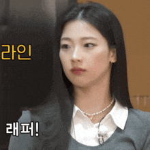 a woman wearing a pearl necklace and a gray sweater looks at the camera with a foreign language behind her