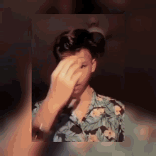 a young man wearing a floral shirt covering his face with his hand
