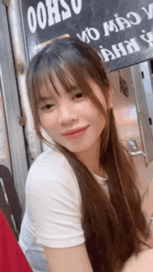 a young woman with long hair and bangs is smiling and looking at the camera while wearing a white shirt .