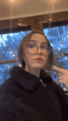 a woman wearing glasses and a black jacket is standing in front of a window