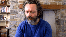 a man with a beard wearing a blue shirt is sitting in front of a brick wall