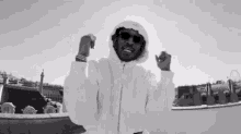 a man in a white hoodie and sunglasses is standing on a rooftop with his arms in the air .