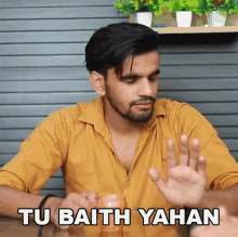 a man in a yellow shirt is sitting at a table with his hand up and the words tu baith yahan written below him