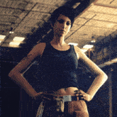 a woman in a black tank top and brown pants stands with her hands on her hips in a dark room