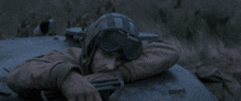 a man wearing a helmet and goggles rests his head on the hood of a car